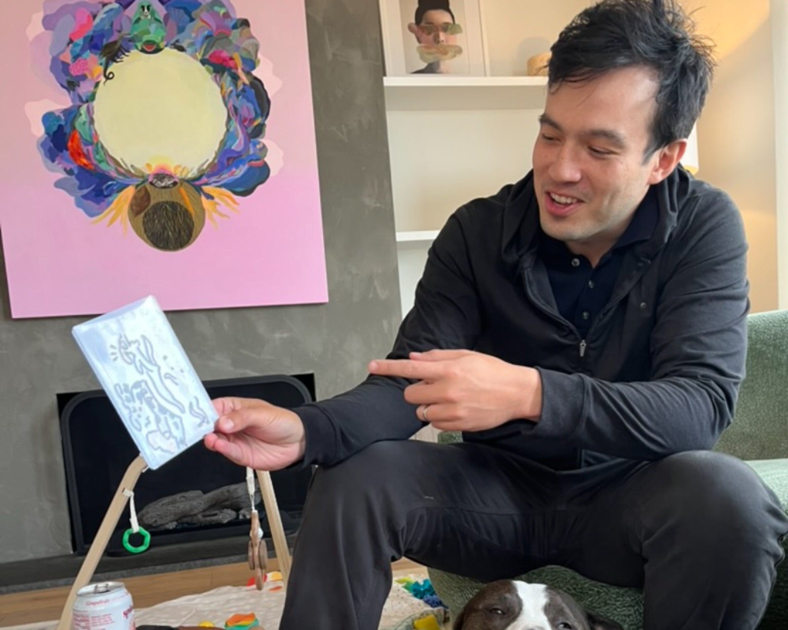 A man at home showing his doodled blob card, his dog resting on the coffee table